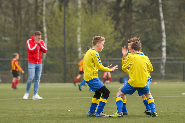 Drie juichende voetballers na het doelpunt en een balende coach van de tegenpartij